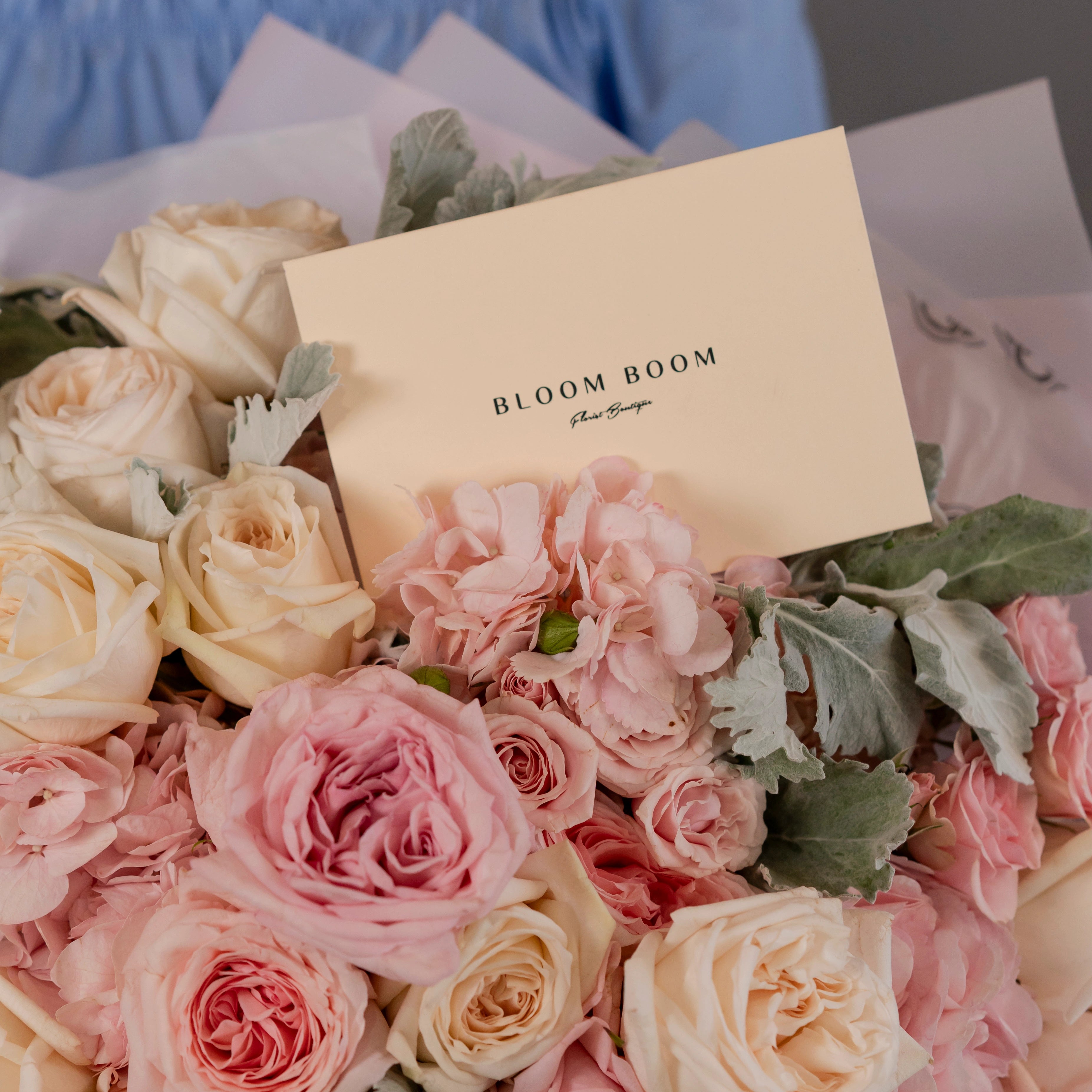 Bloom Boom Flowers branded bouquet with blush pink and cream roses, dusty miller leaves in soft pink wrapping
