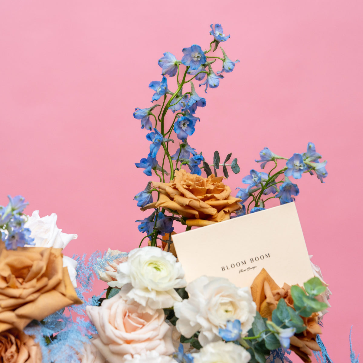 Mixed flower bouquet with blue delphiniums, peach roses, white ranunculus on pink background with Bloom Boom card