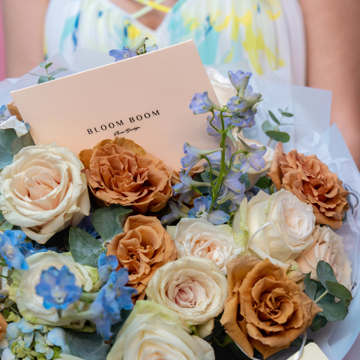Mixed rose bouquet with cream, peach and brown roses, blue delphinium flowers, eucalyptus leaves and Bloom Boom card
