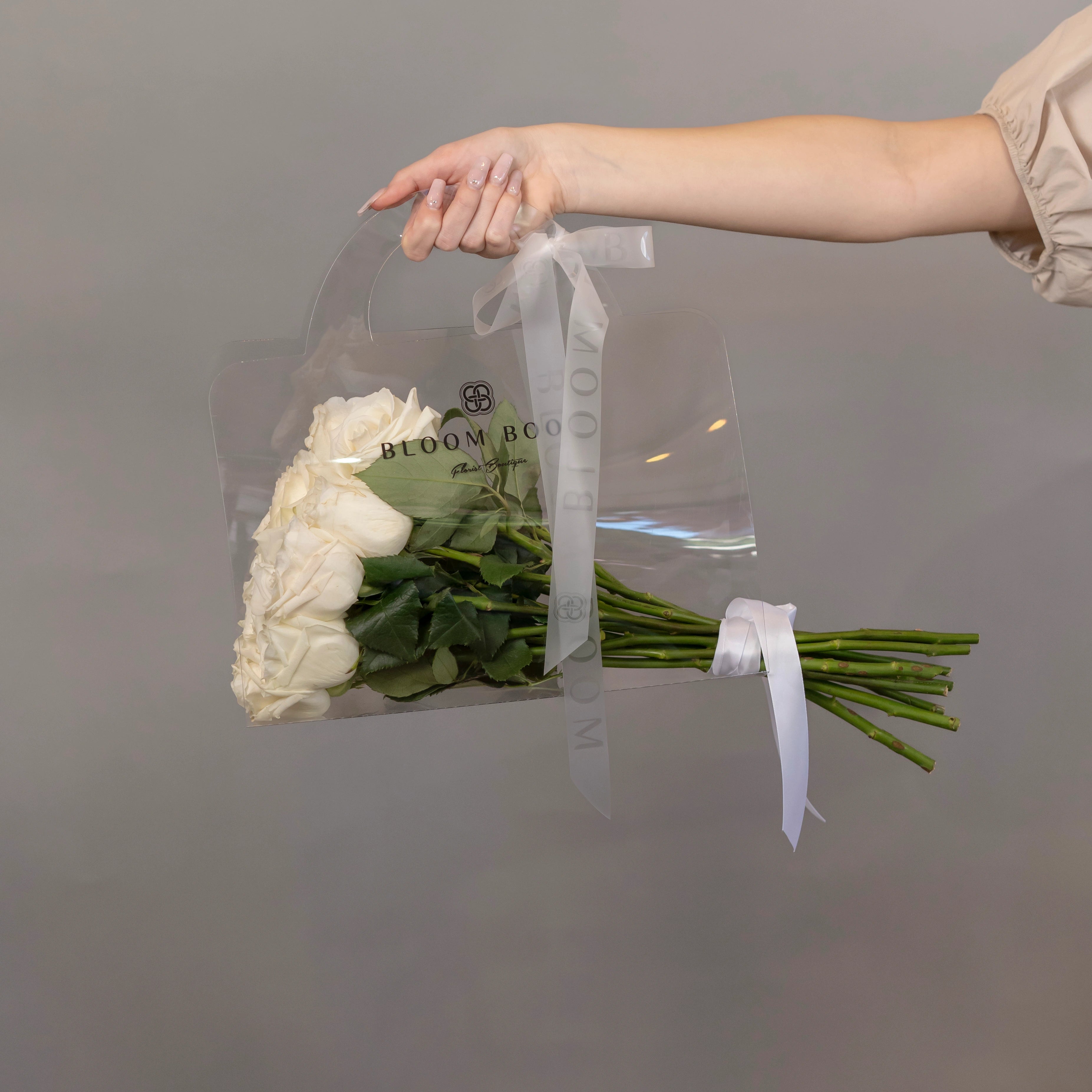 Clear acrylic flower box with white roses and green foliage, branded Bloom Boom, tied with white satin ribbon
