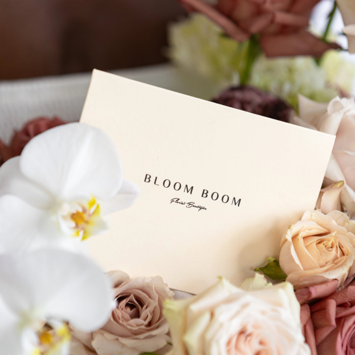 Bloom Boom cream business card displayed with white orchid, peach roses, and soft pink flowers in gift arrangement
