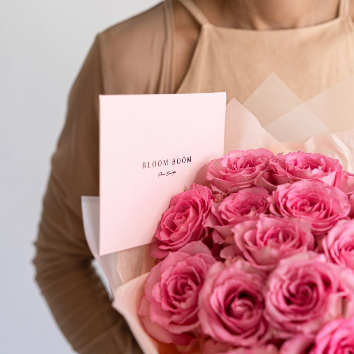 Pink roses bouquet in cream paper wrapping with Bloom Boom branded card, held by person in beige clothing