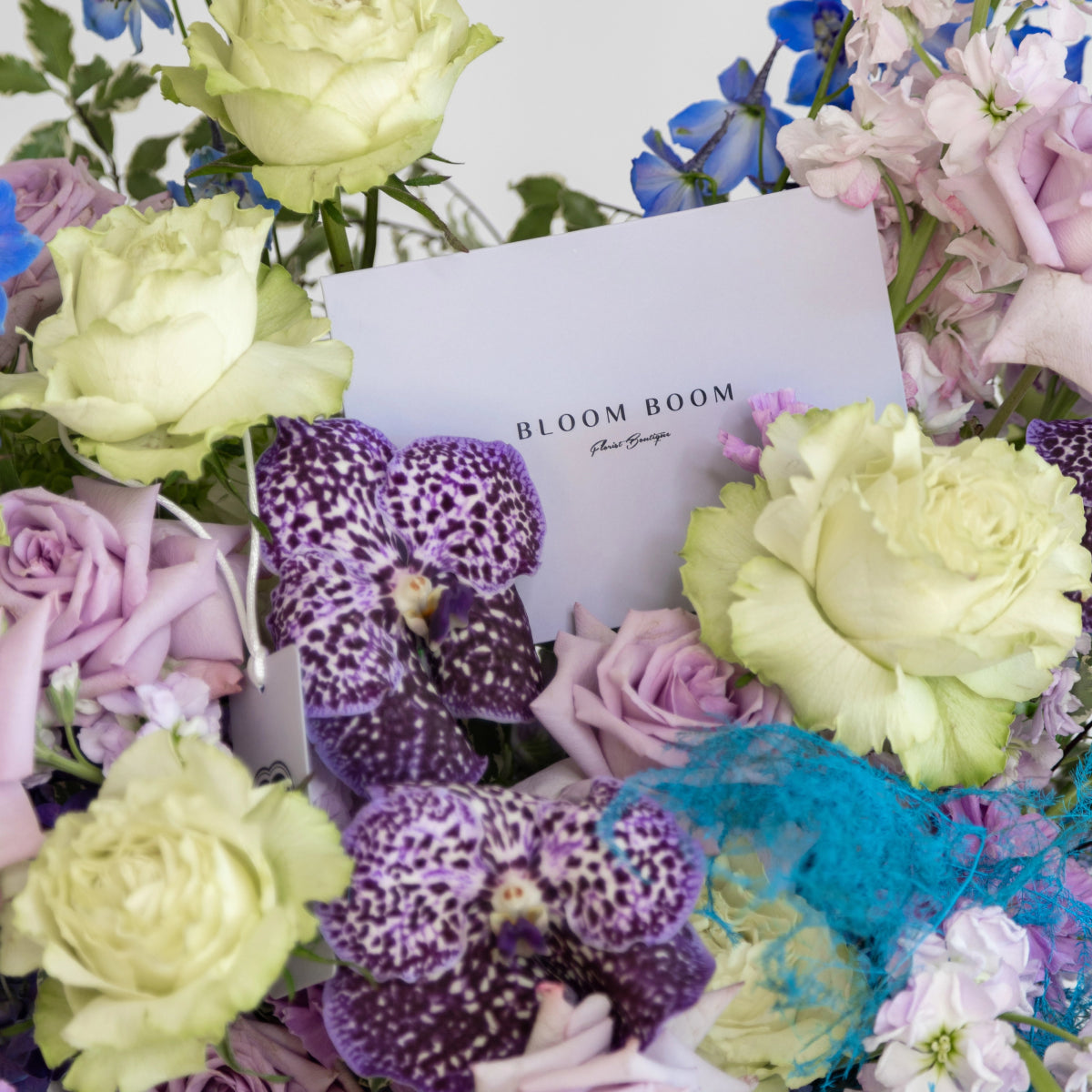 Bloom Boom flower arrangement with purple spotted orchids, cream roses, and pink flowers with branded white card