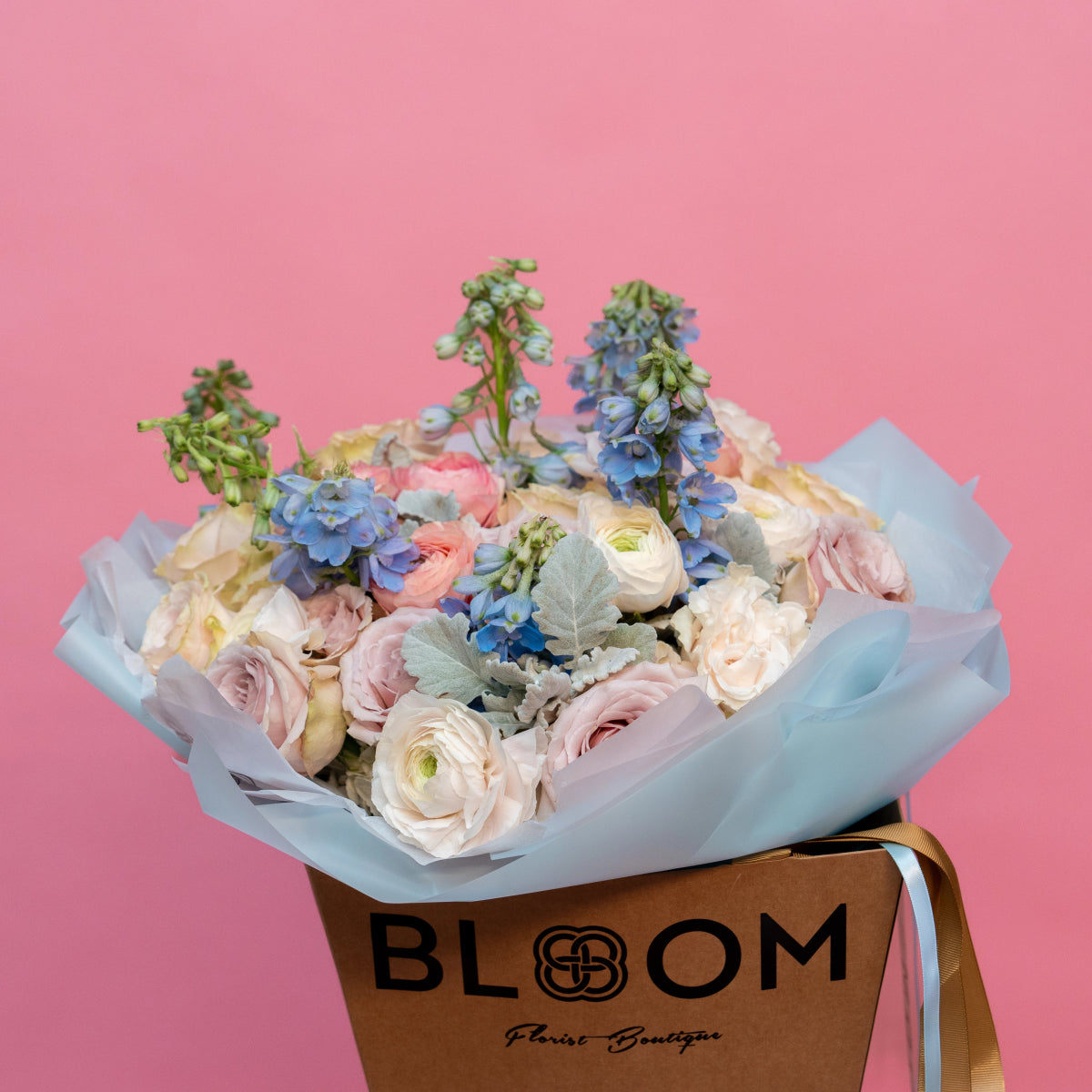 Mixed flower bouquet with white ranunculus, pink roses, blue delphiniums in light blue tissue paper wrapping