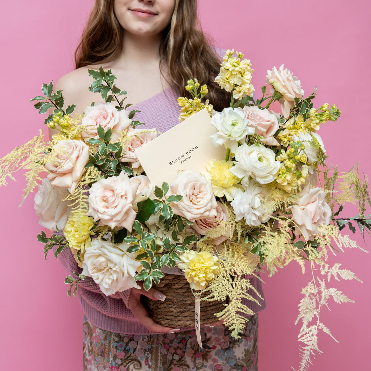 Large wicker basket filled with blush pink roses, white ranunculus, yellow flowers and golden ferns on pink background