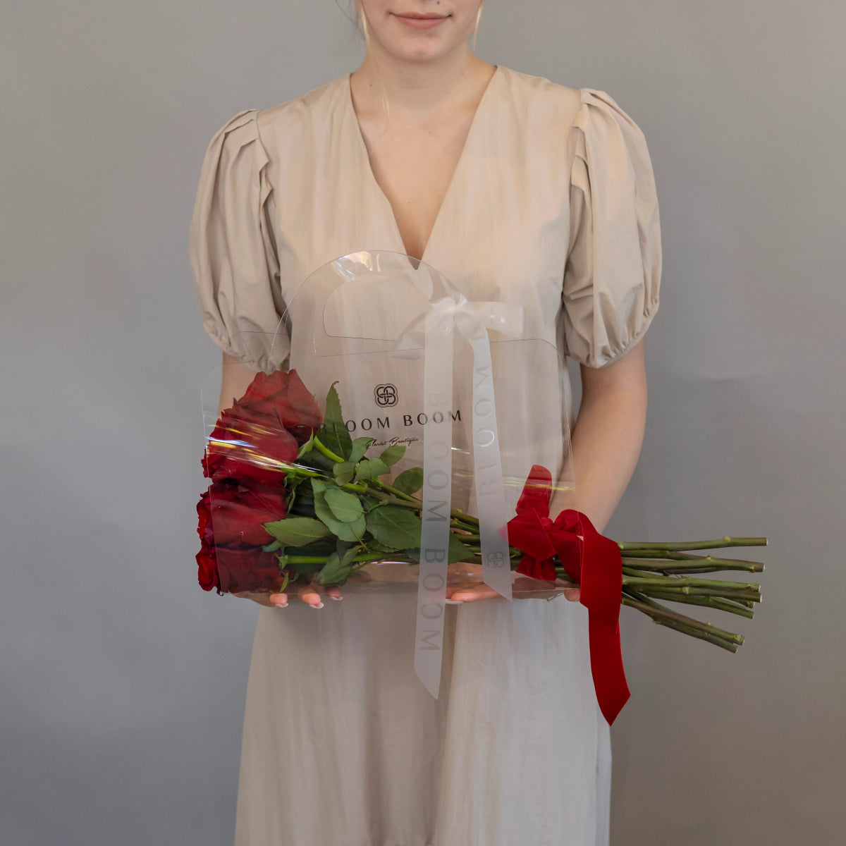 Clear acrylic flower box with 12 red roses and red ribbon bow held by woman in beige dress