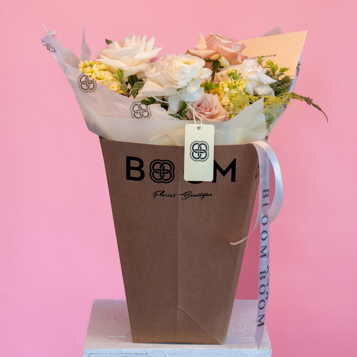 White and pink roses with yellow flowers in brown BOOM Florist Boutique paper bag with ribbon on pink background