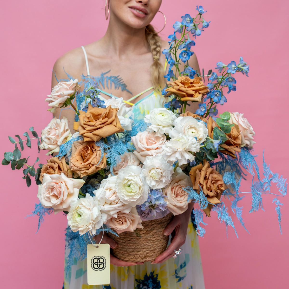 Wicker flower basket with peach roses, white ranunculus, blue delphiniums and eucalyptus on pink background