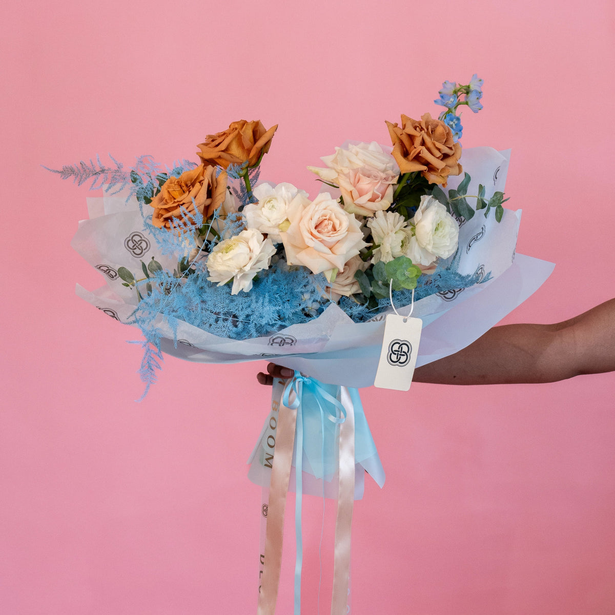 Mixed flower bouquet with peach roses, white ranunculus, blue dried flowers in tissue paper wrapping with ribbon