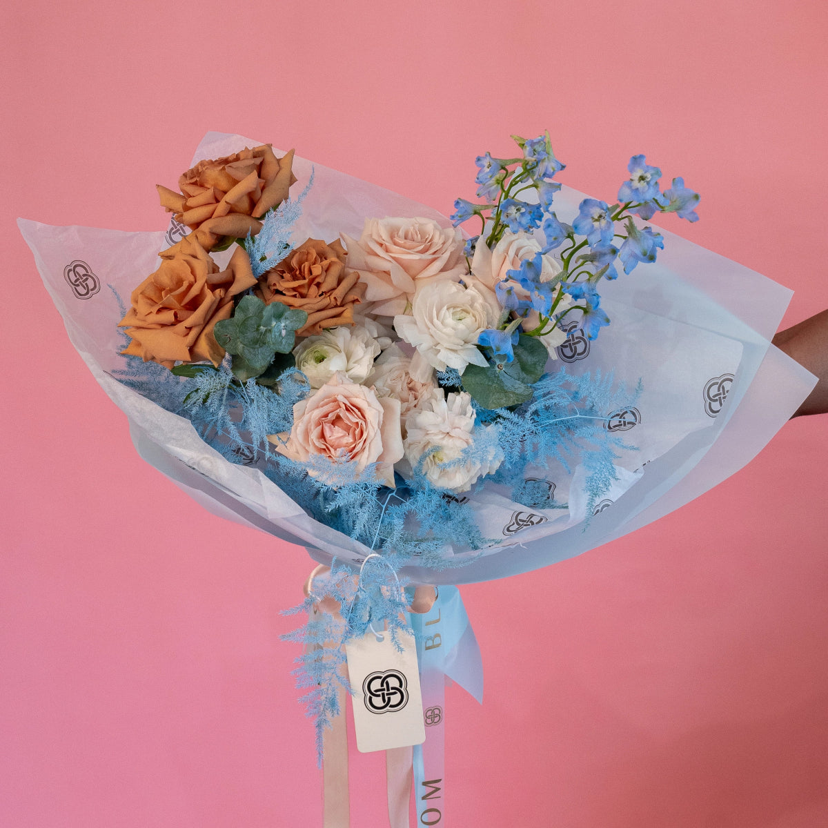 Mixed flower bouquet with orange roses, cream ranunculus, blue delphinium in clear wrapping with blue ribbon