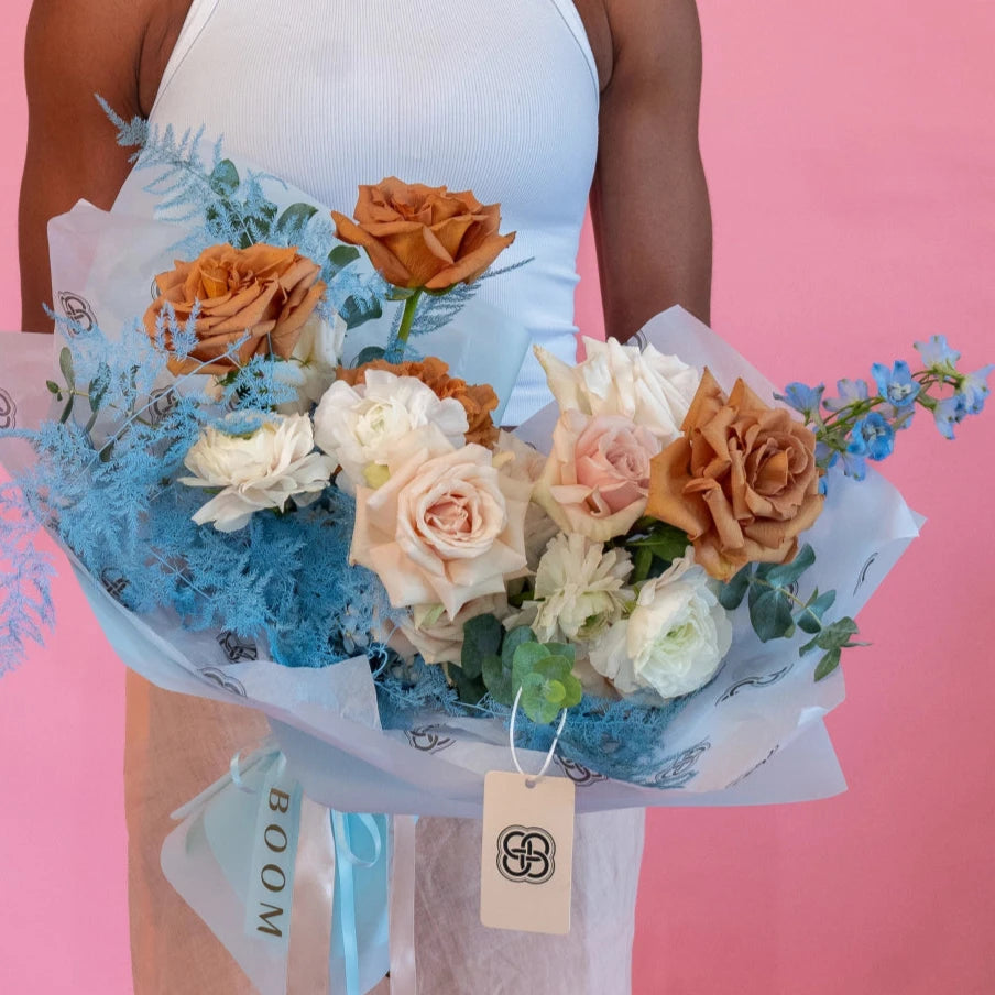 Mixed flower bouquet with orange roses, white ranunculus, pink roses and blue dried flowers in white paper wrapping