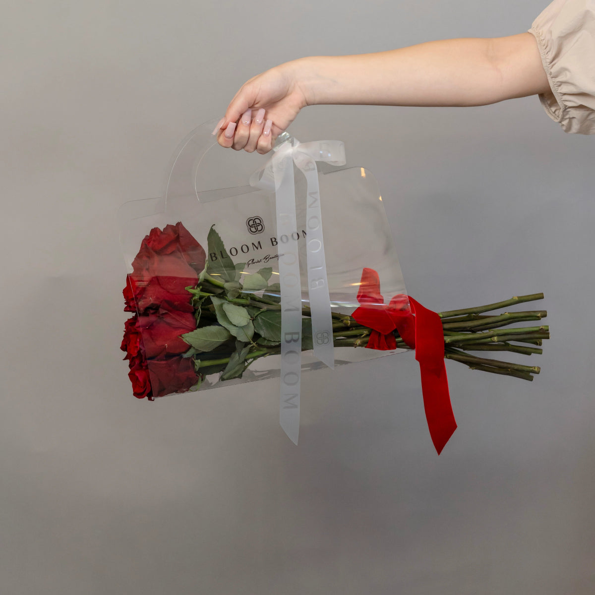 Clear acrylic flower carrier box holding 12 red roses with green stems and red ribbon, carried by hand