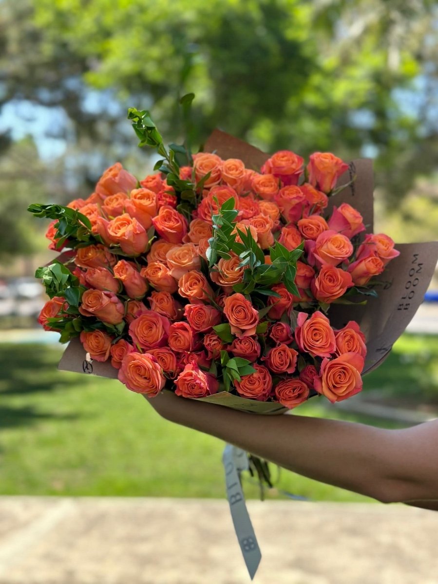 Large bouquet of vibrant coral and orange roses with green foliage wrapped in brown kraft paper outdoors