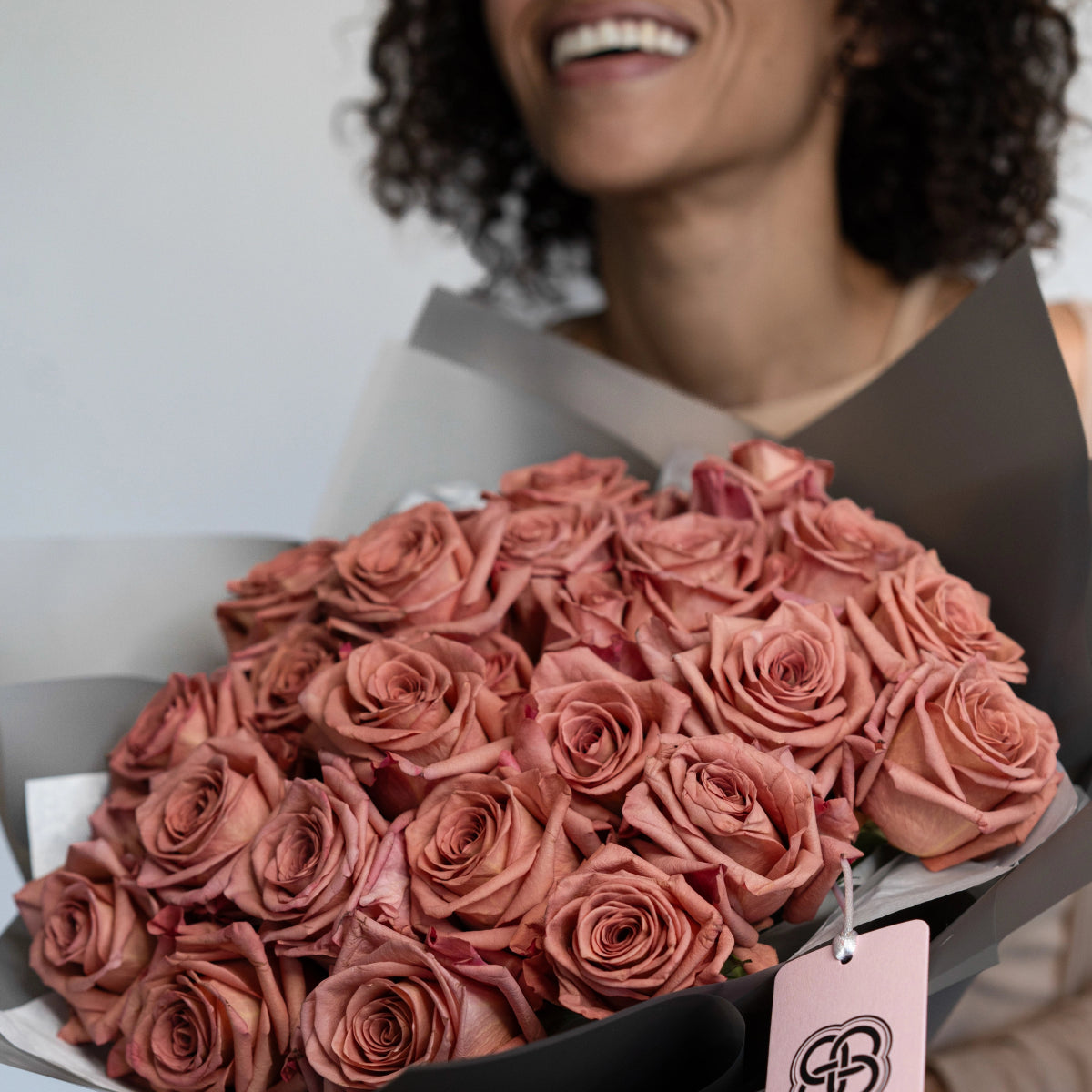 Dusty rose pink roses bouquet wrapped in black paper with white tag held by smiling woman