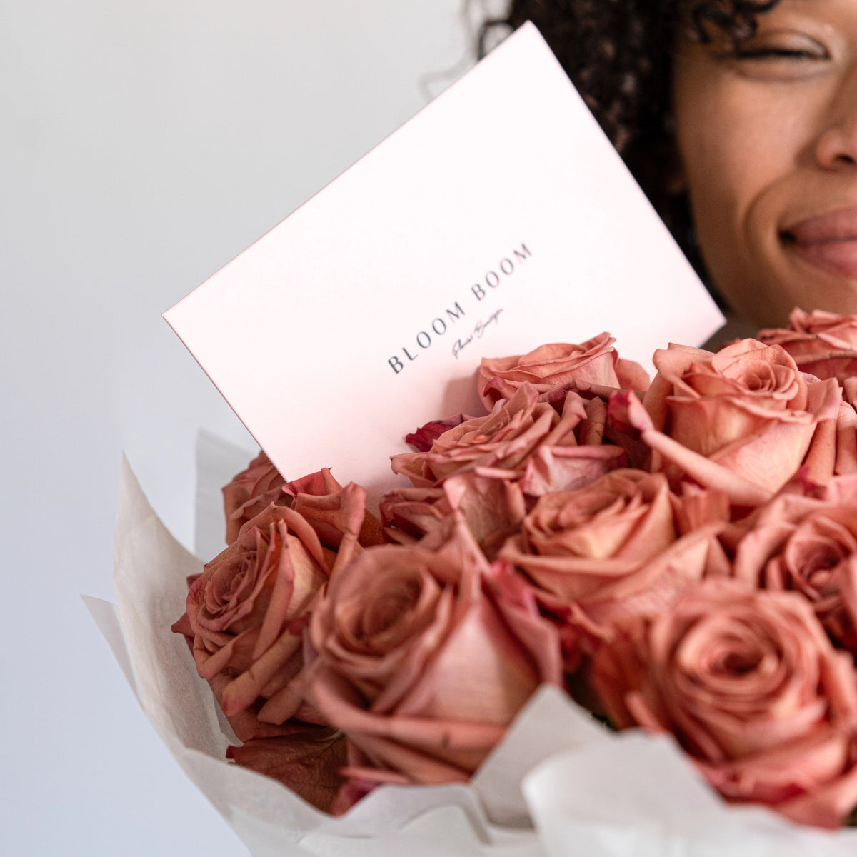 Dusty pink roses bouquet wrapped in white paper with Bloom Boom brand card held by smiling woman