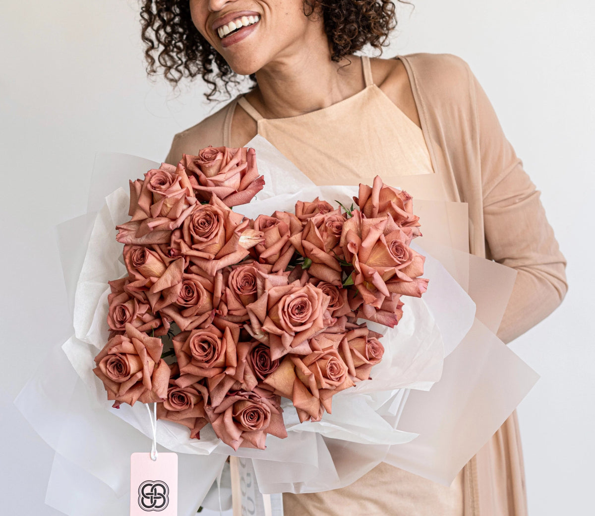 Woman holding large bouquet of dusty pink roses wrapped in white paper with brand tag