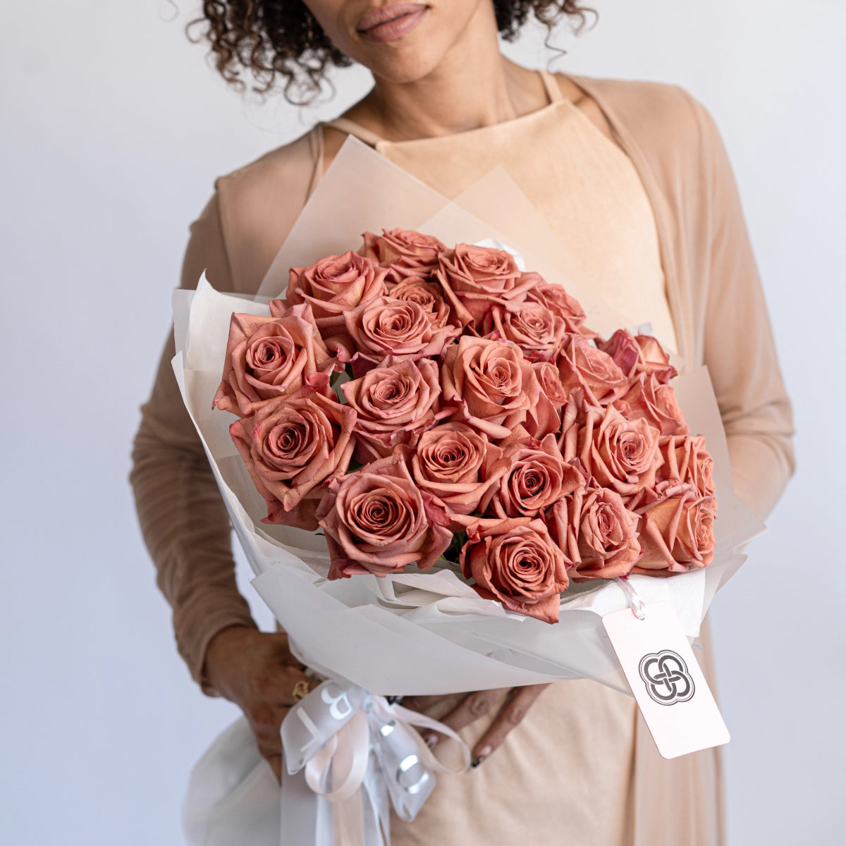 Woman holding dusty rose pink roses bouquet wrapped in white paper with ribbon and brand tag