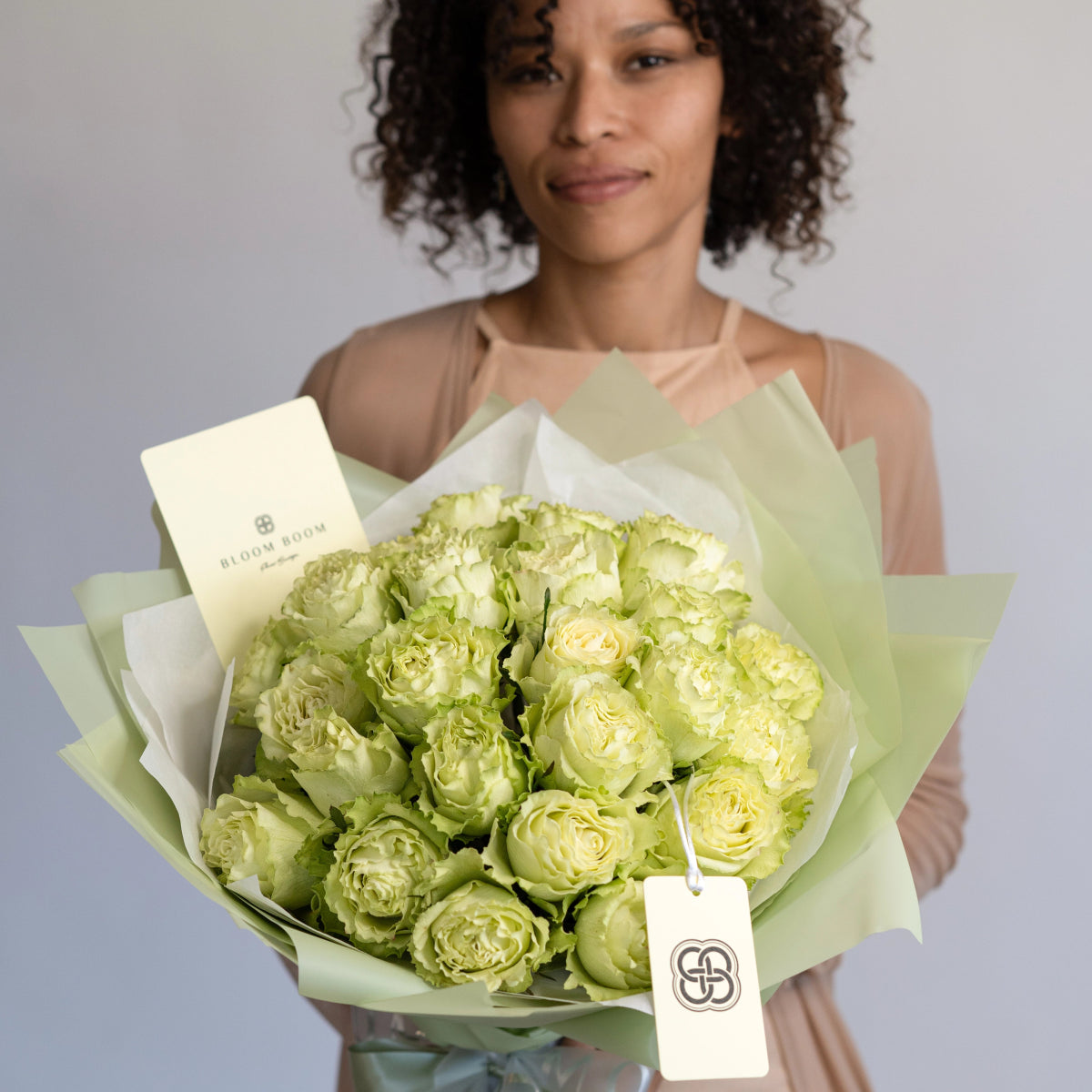 Woman holding bouquet of pale green ruffled roses wrapped in sage green paper with Bloom Room gift tag