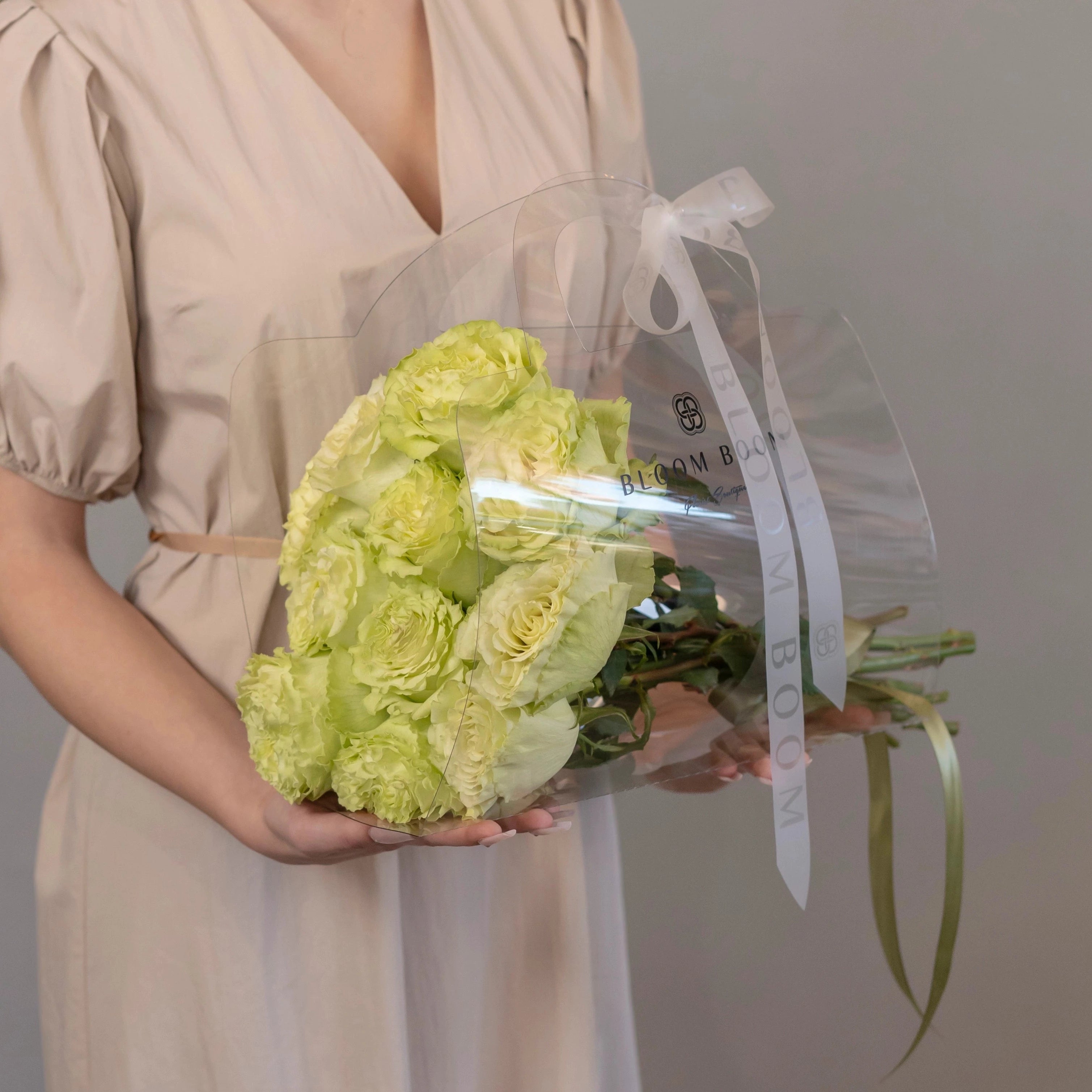 Lime green and cream rose bouquet wrapped in clear cellophane with white ribbon held by woman in beige dress