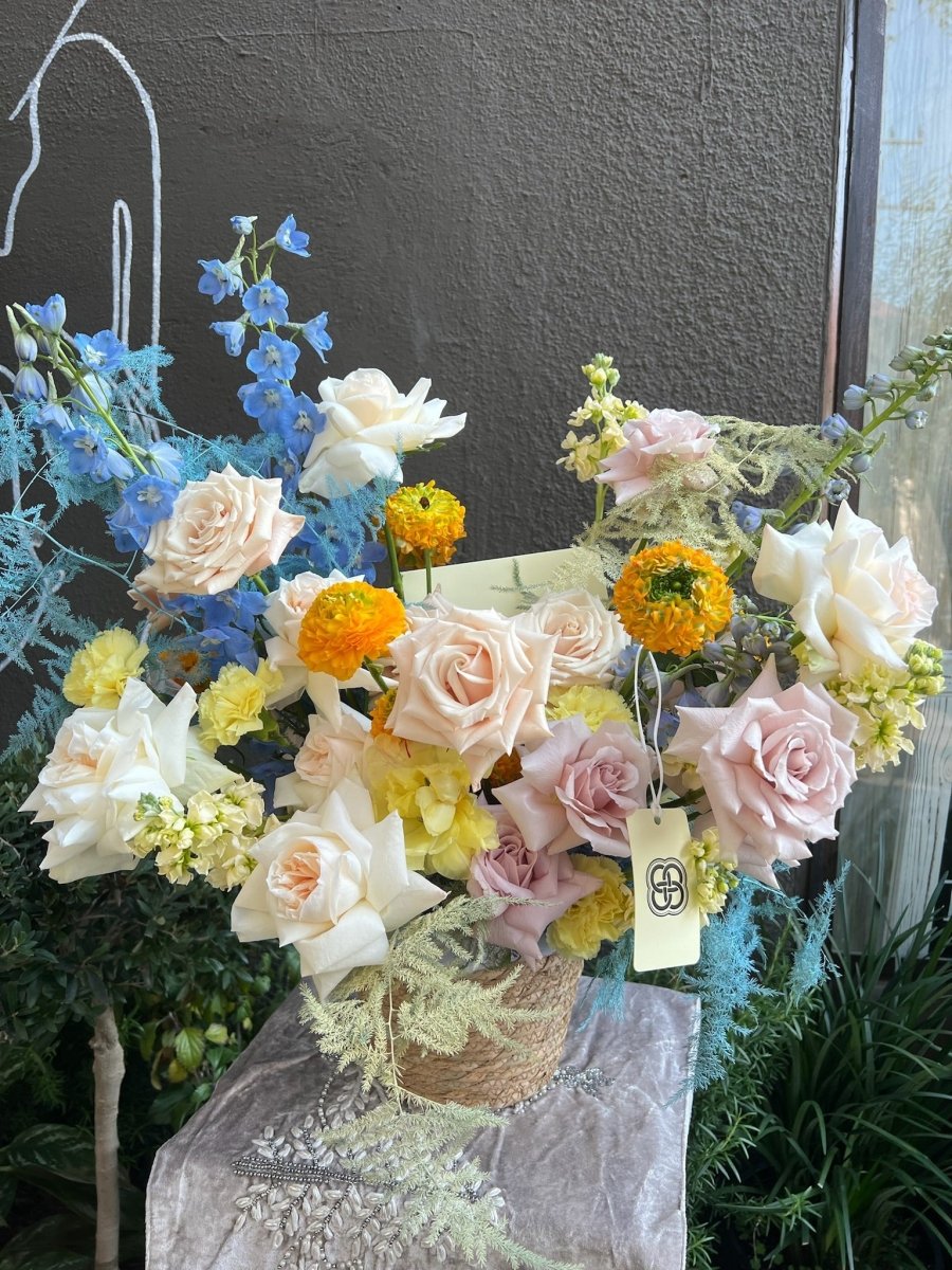 Mixed flower basket with blush roses, white tulips, blue delphiniums, orange marigolds in woven natural basket