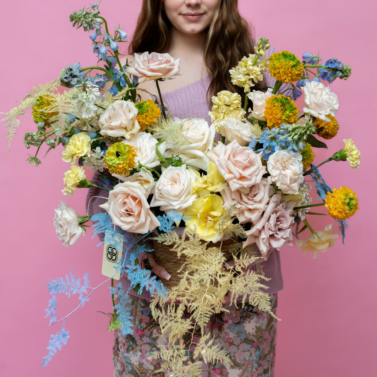 Large mixed flower bouquet with blush roses, yellow carnations, blue dusty miller in woven basket against pink backdrop
