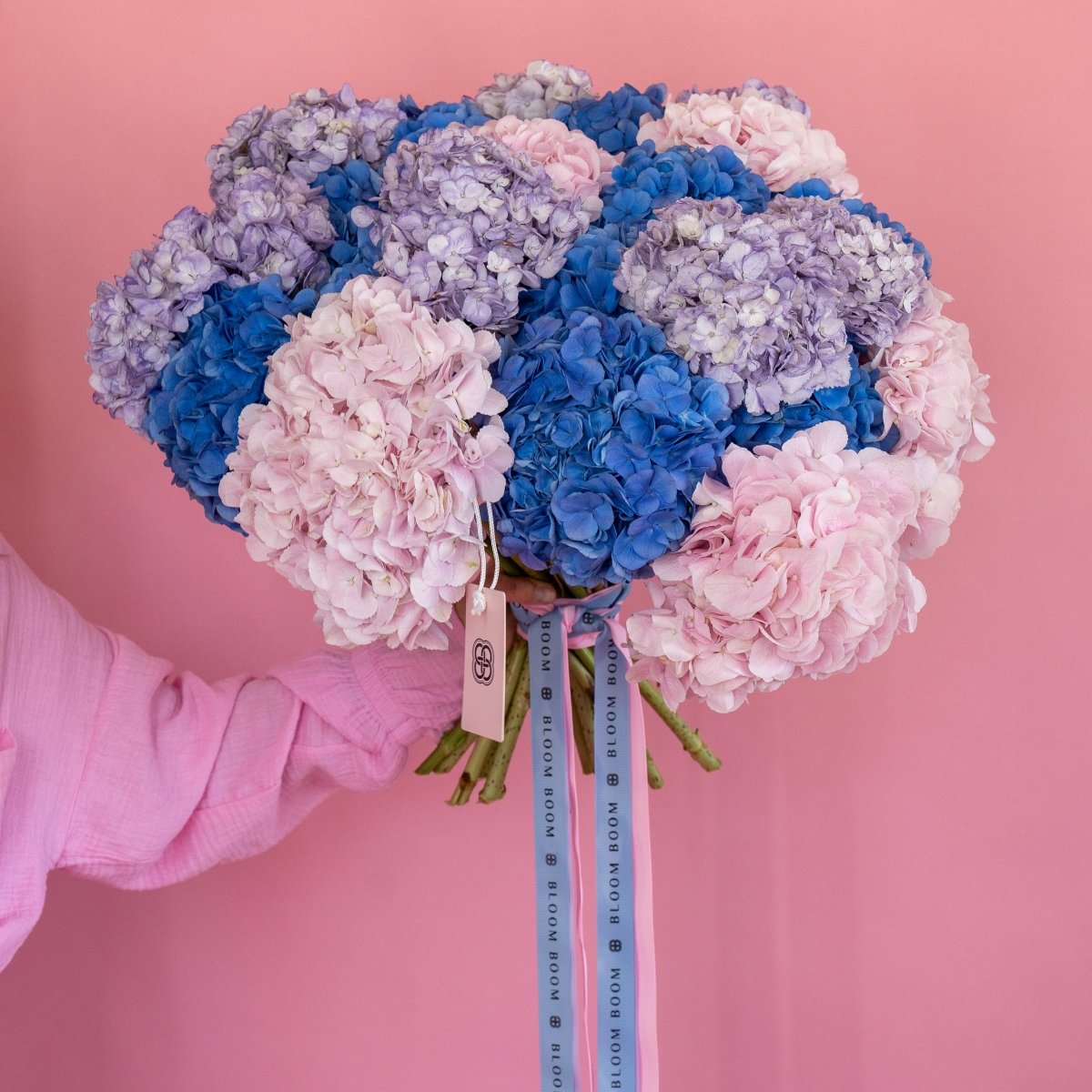 Mixed hydrangea bouquet with blue, pink, and purple blooms tied with branded ribbons on coral background