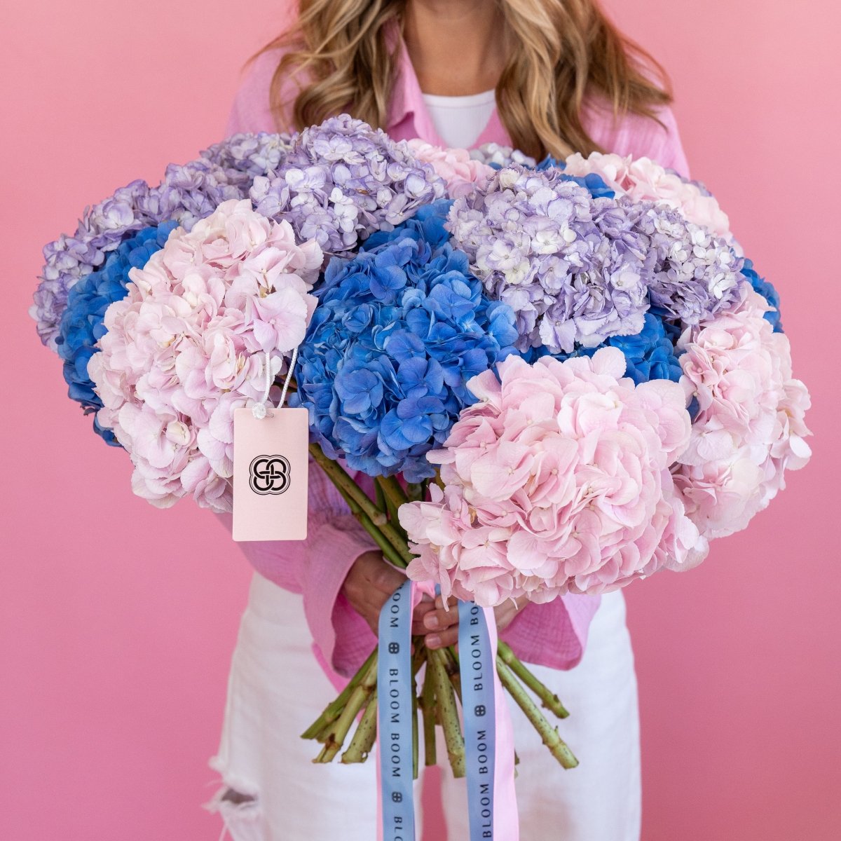 Large mixed hydrangea bouquet with pink, blue, and purple blooms tied with branded ribbon on pink background