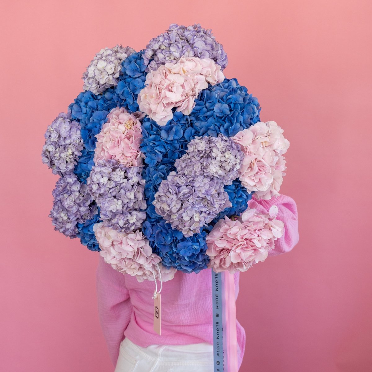 Mixed hydrangea bouquet with blue, pink, and purple blooms in pink gift box with ribbon on coral background
