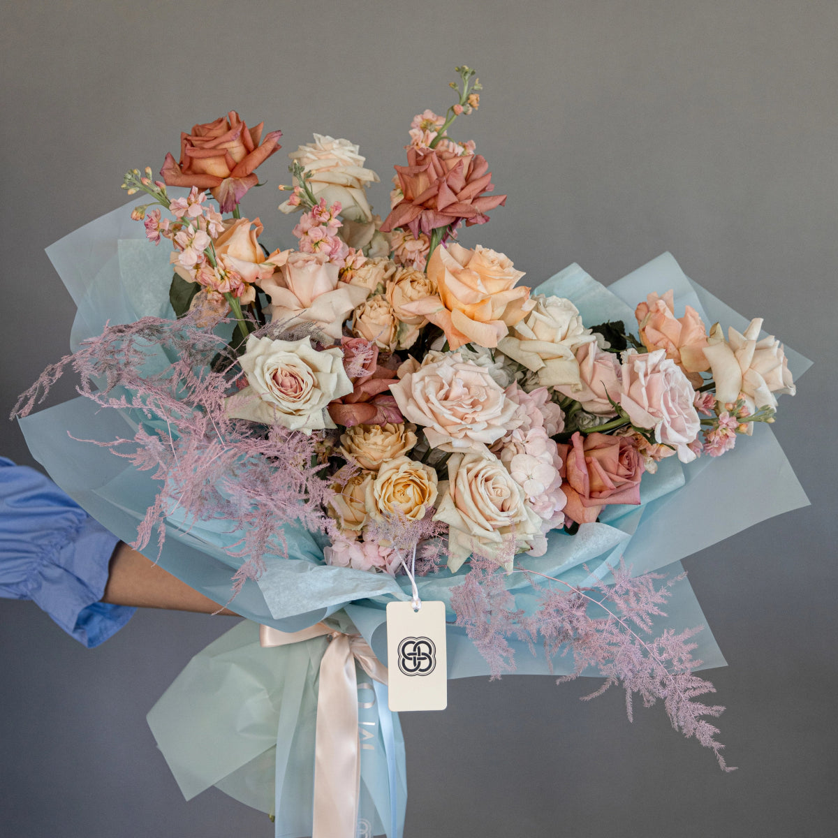 Mixed rose bouquet in peach, cream and dusty pink tones with blue tissue paper wrapping and brand tag