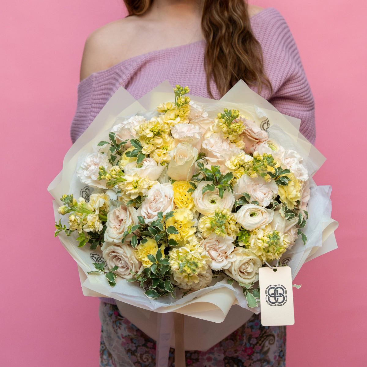 Pastel bouquet with cream roses, yellow ranunculus and green foliage wrapped in white paper against pink background