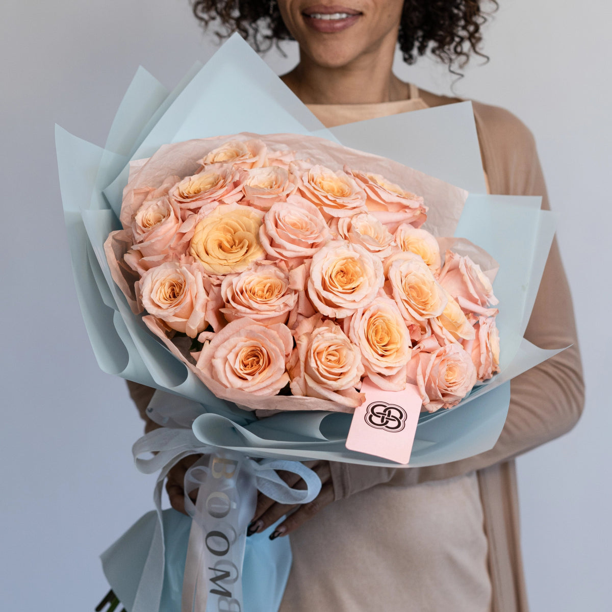 Elegant bouquet of 25 peach and coral roses wrapped in soft blue paper with ribbon bow and brand tag