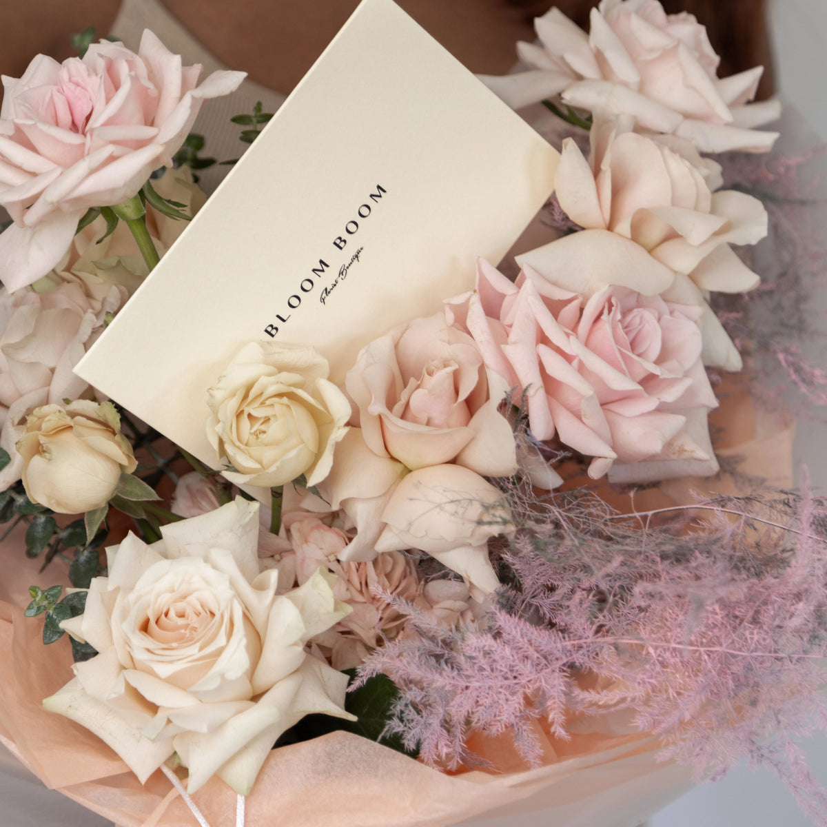 Elegant pink and cream rose bouquet with white card reading 'BLOOM BOOM' wrapped in soft pink paper