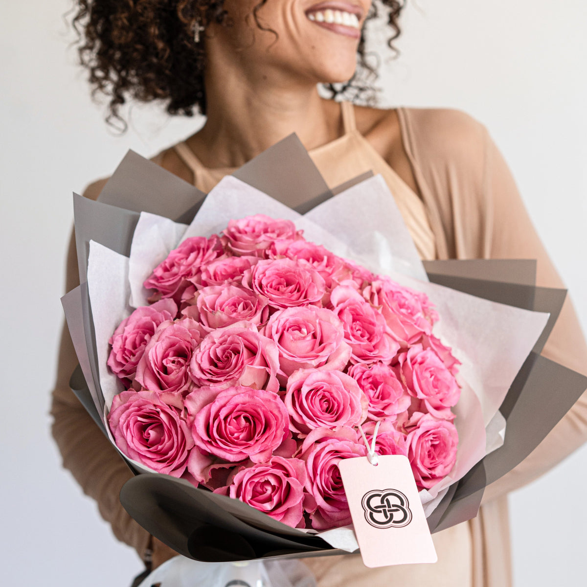 Luxurious pink rose bouquet wrapped in white and gray paper with elegant gift tag held by smiling woman