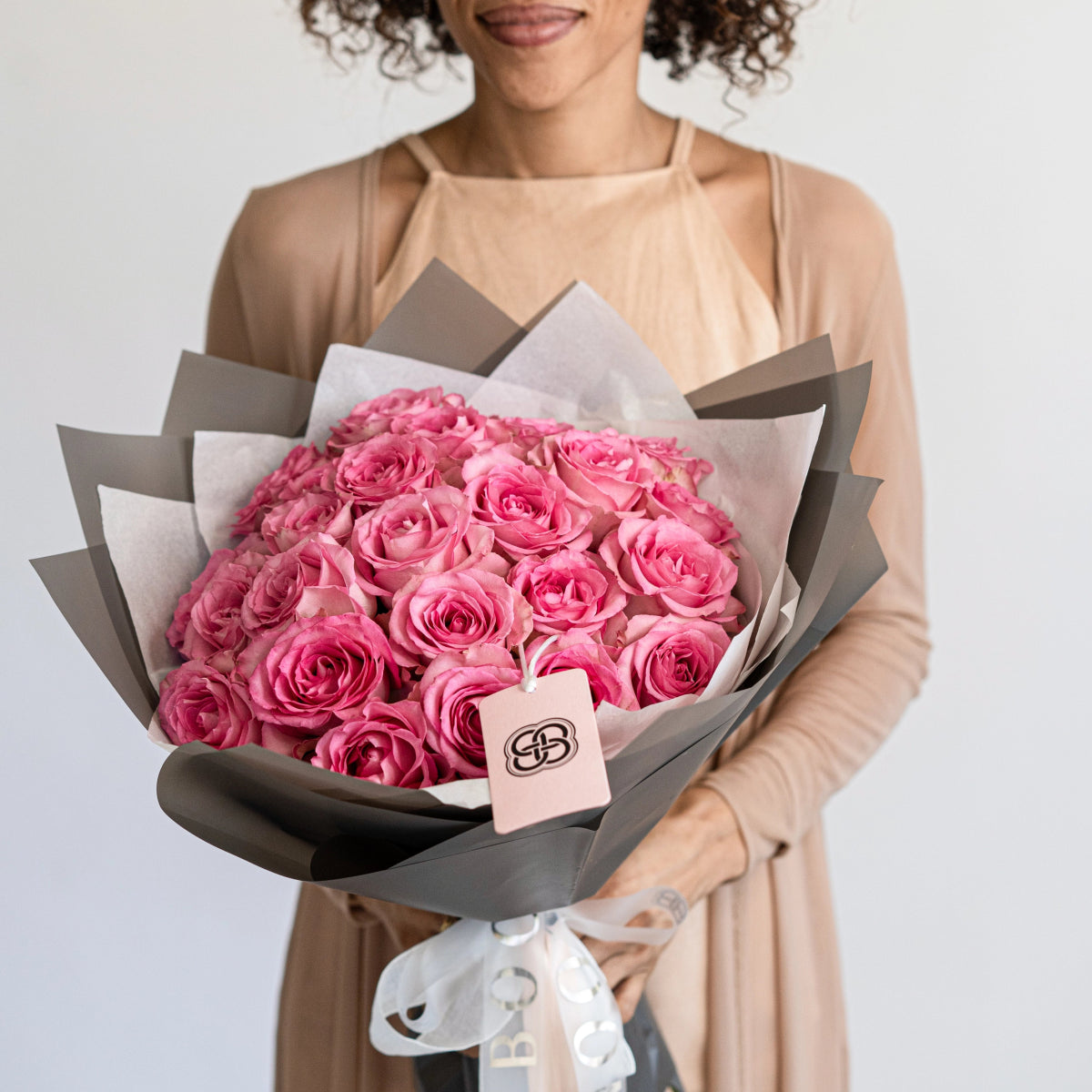 Large bouquet of 25 pink roses wrapped in gray and white paper with brand tag, held by woman in beige dress