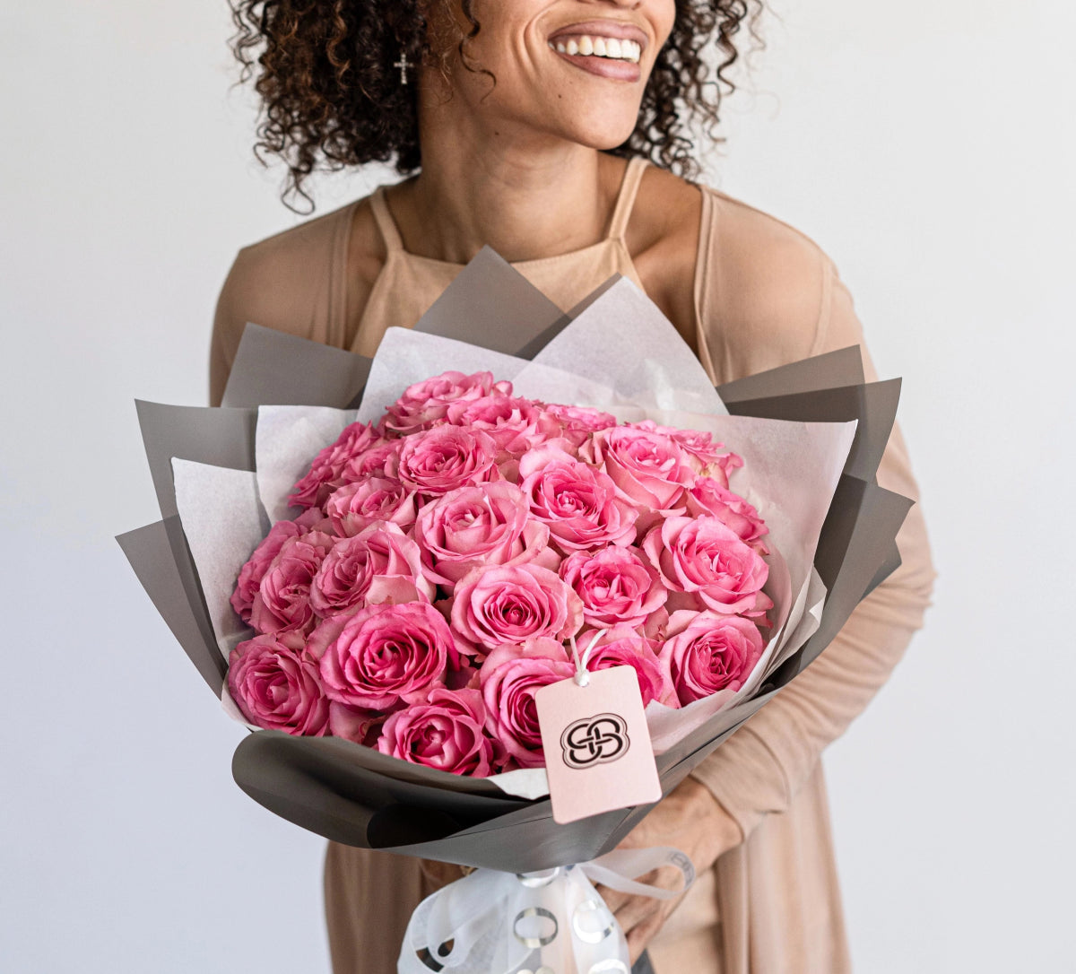 Pink rose bouquet with gray and white wrapping paper held by smiling woman in beige outfit