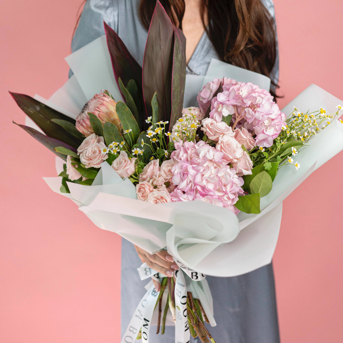 Pink rose and hydrangea bouquet with white chamomile flowers wrapped in sage green paper on coral background