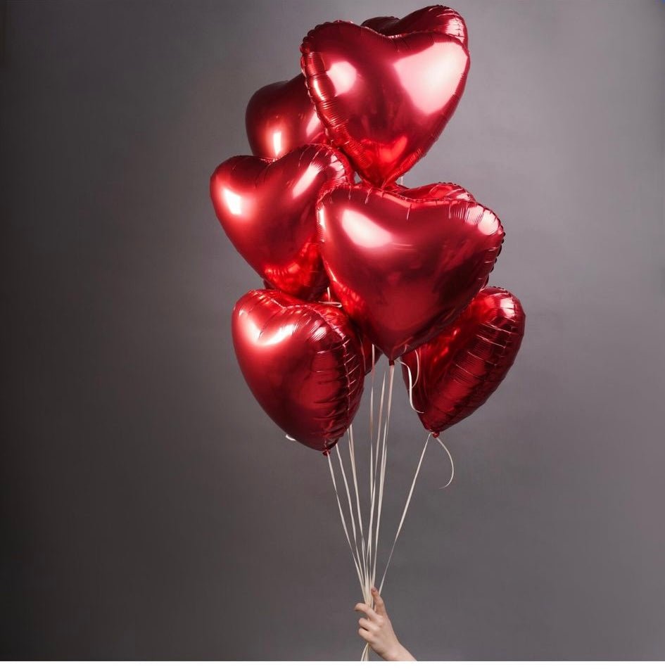 Red metallic heart-shaped foil balloons bunch with white ribbons held by hand against gray background