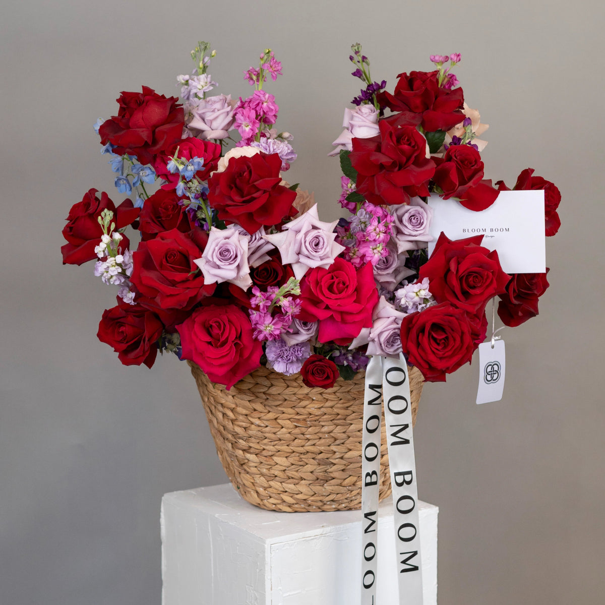 Mixed rose bouquet in woven basket with red and lavender roses, purple stock flowers, and Bloom Boom ribbon