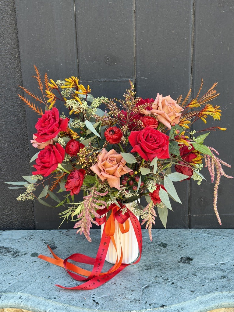 Red October fall arrangement of red roses, toffee roses, red ranunculus, kangaroo paw and pink amaranthus in a fluted white vase with red and orange ribbons