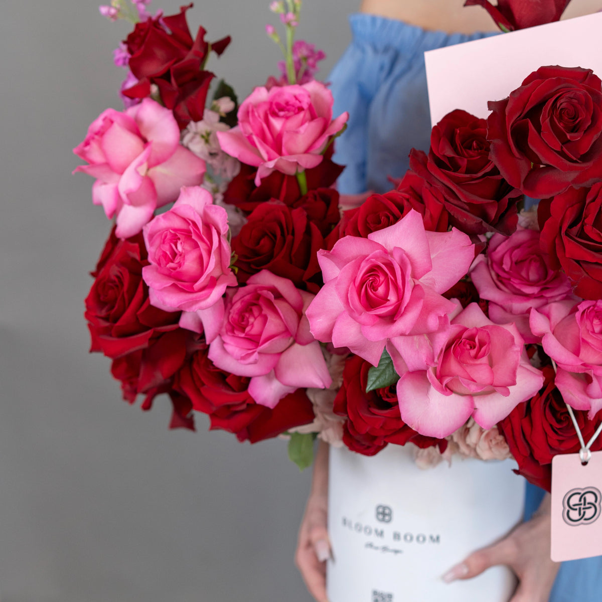 White round flower box filled with deep red and soft pink roses, held by person in blue denim shirt