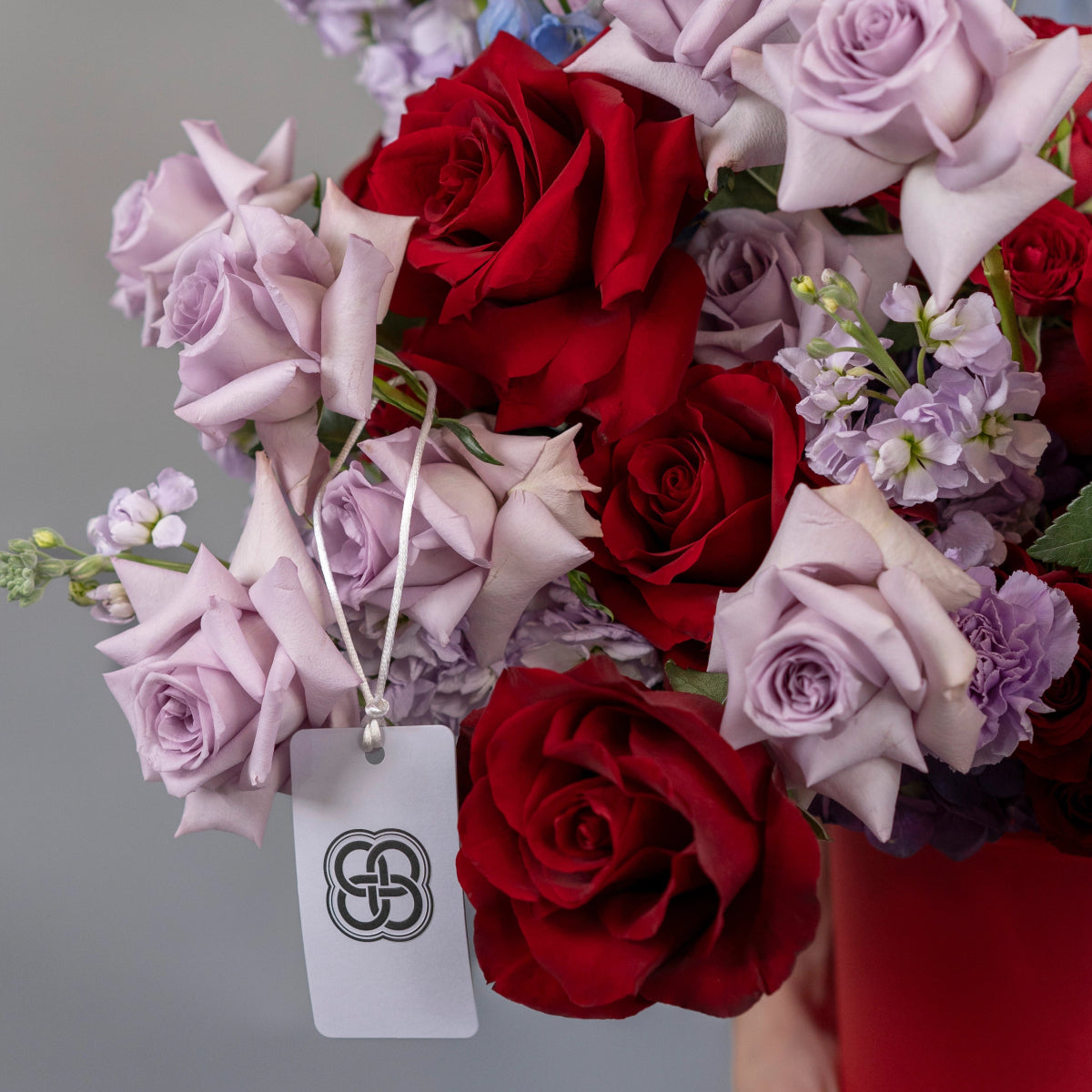 Red roses and lavender purple roses with white stock flowers in burgundy box with branded gift tag
