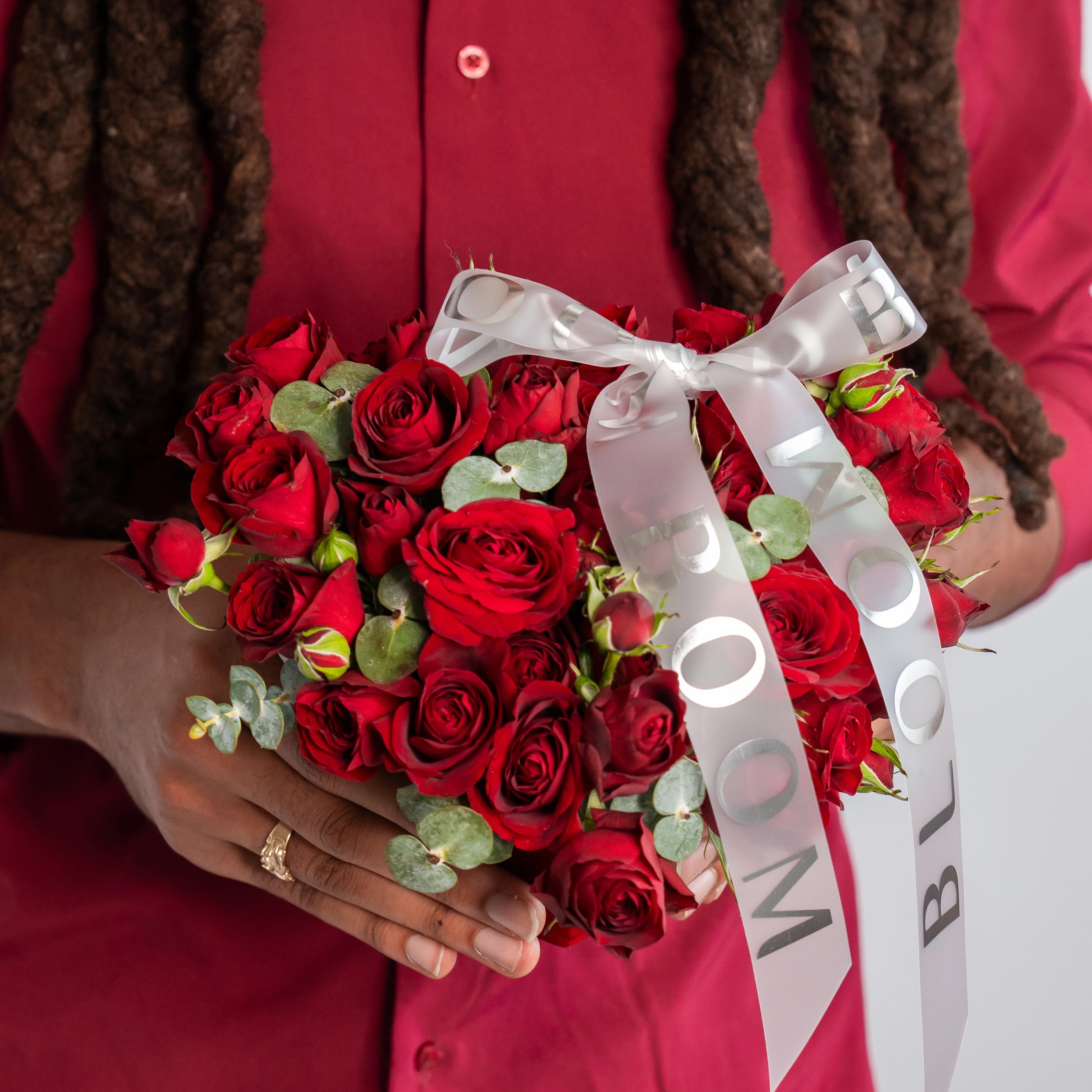 Red rose bouquet with eucalyptus greenery and white satin Bloom Boom ribbon held by person in red dress