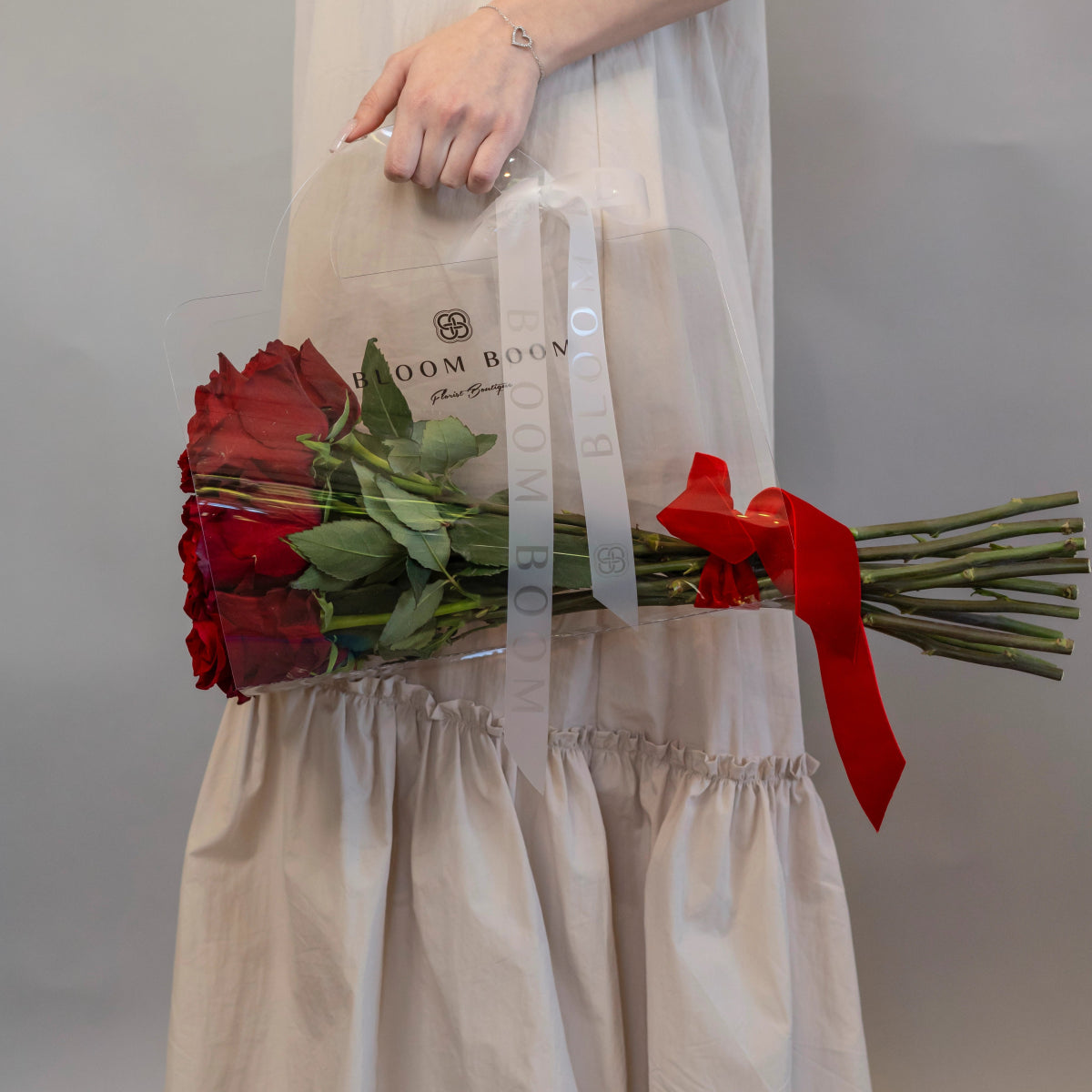 Woman holding dozen red roses with green stems tied with red ribbon in clear Bloom Boom branded bag