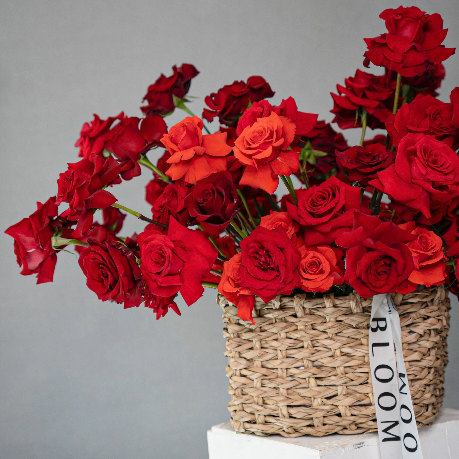 Red roses arrangement in natural woven basket with coral accent blooms and green stems on white surface