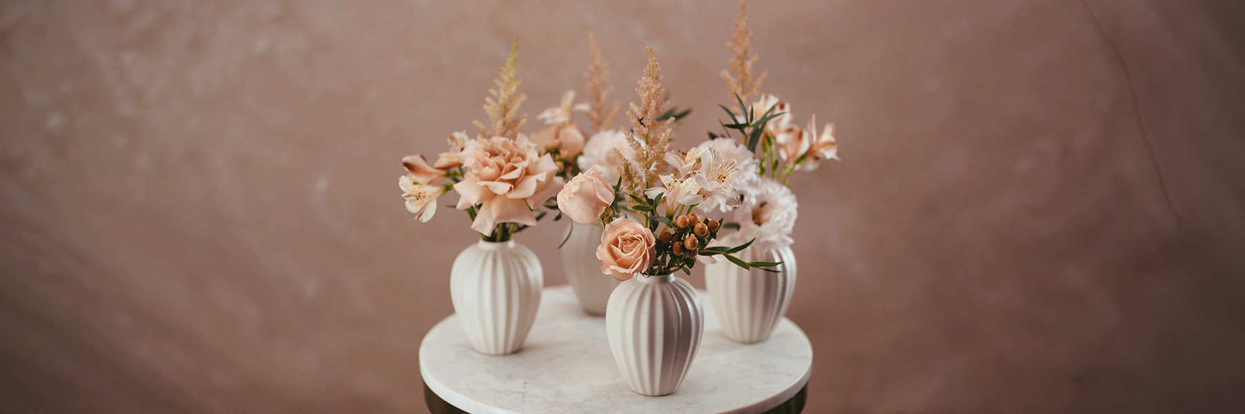 Three ribbed cream ceramic vases with peach roses, white flowers and dried pampas grass on white marble table