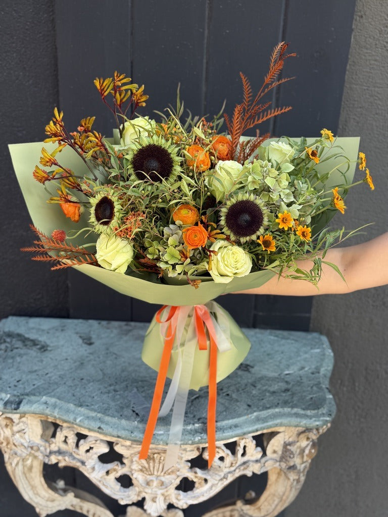 Sunflower Spice fall bouquet of sunflowers, orange ranunculus, pale green roses, hydrangea and kangaroo paw in sage green wrap with orange ribbons
