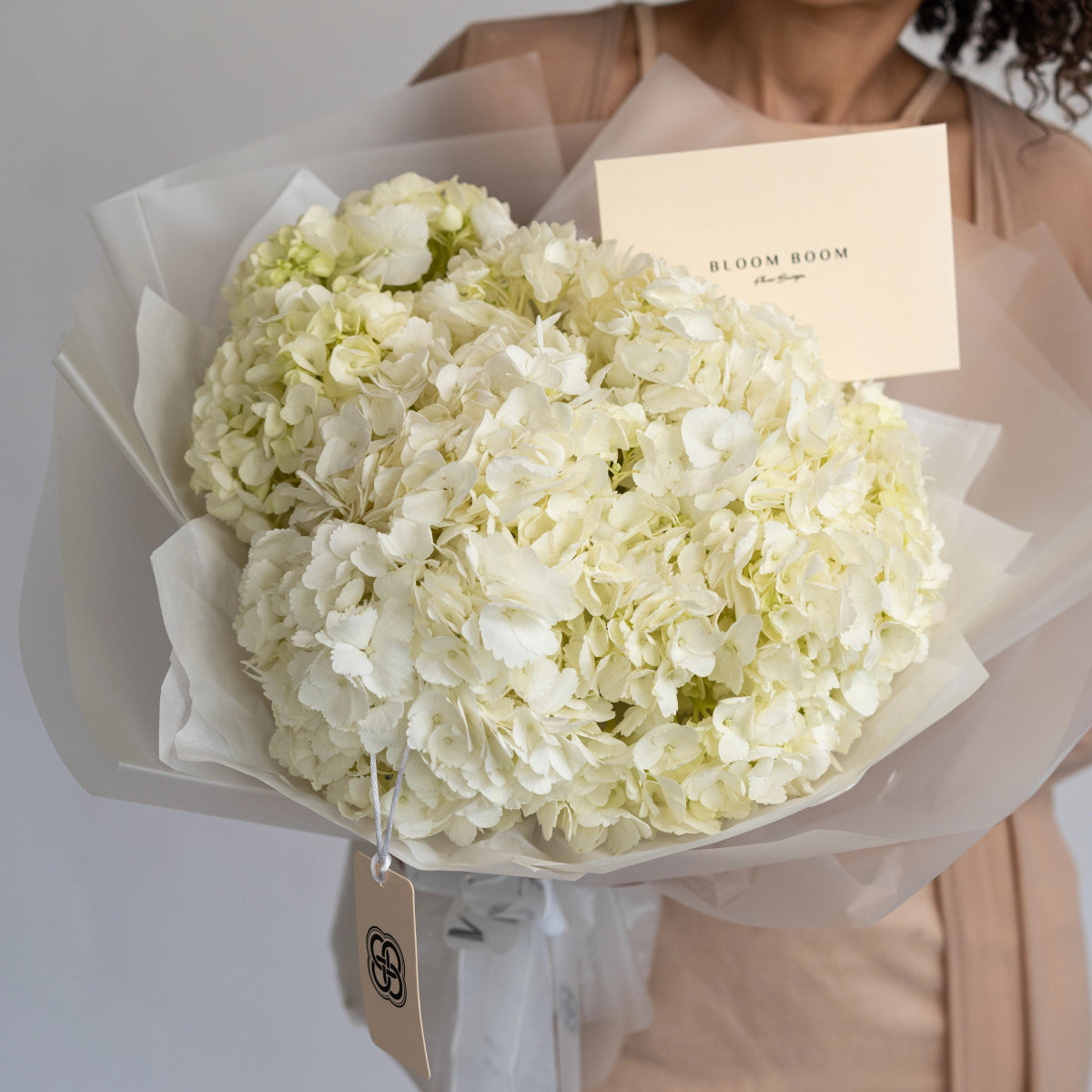 White hydrangea bouquet wrapped in cream tissue paper with Bloom Boom card held by woman in beige dress