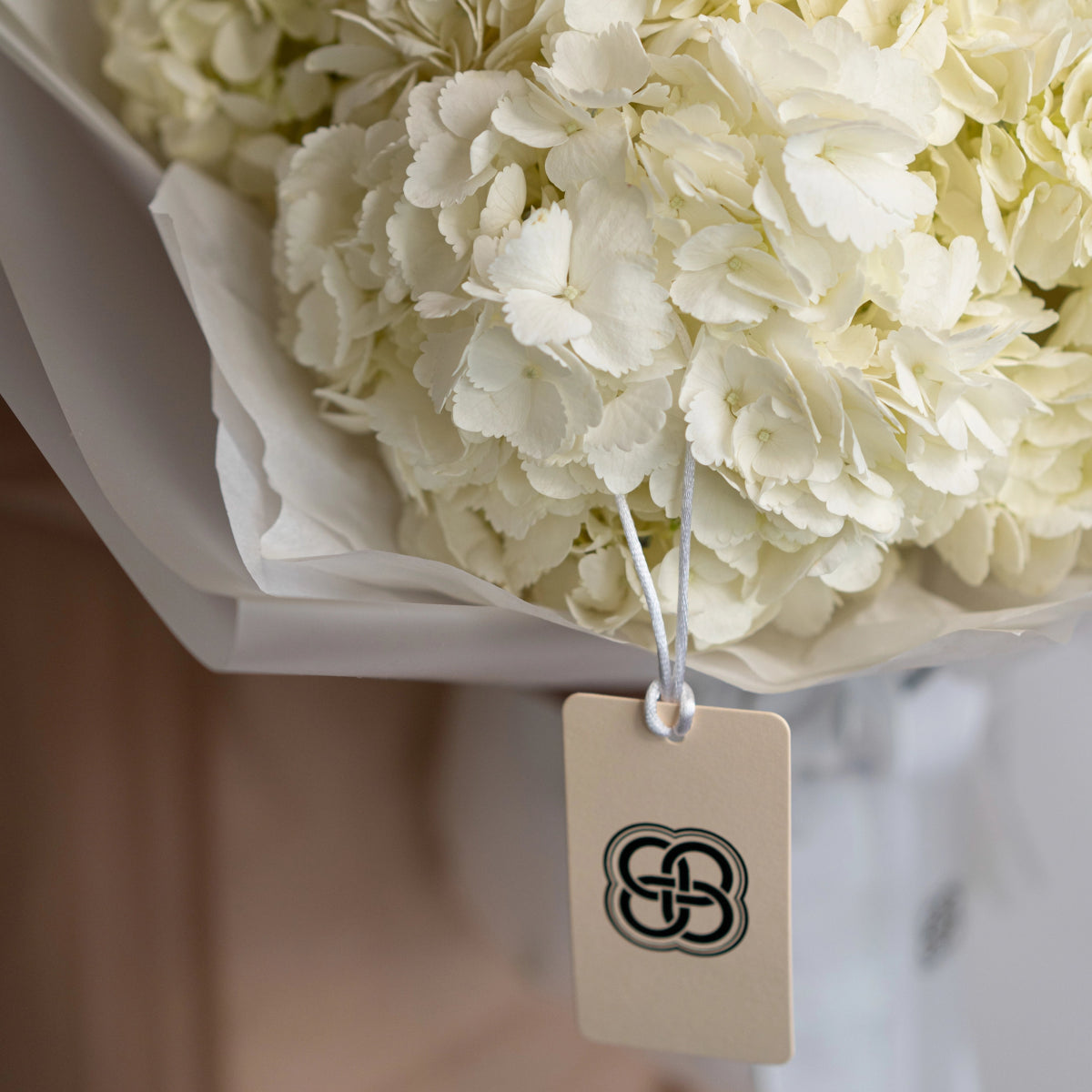 Cream white hydrangea bouquet wrapped in white tissue paper with beige gift tag featuring circular logo