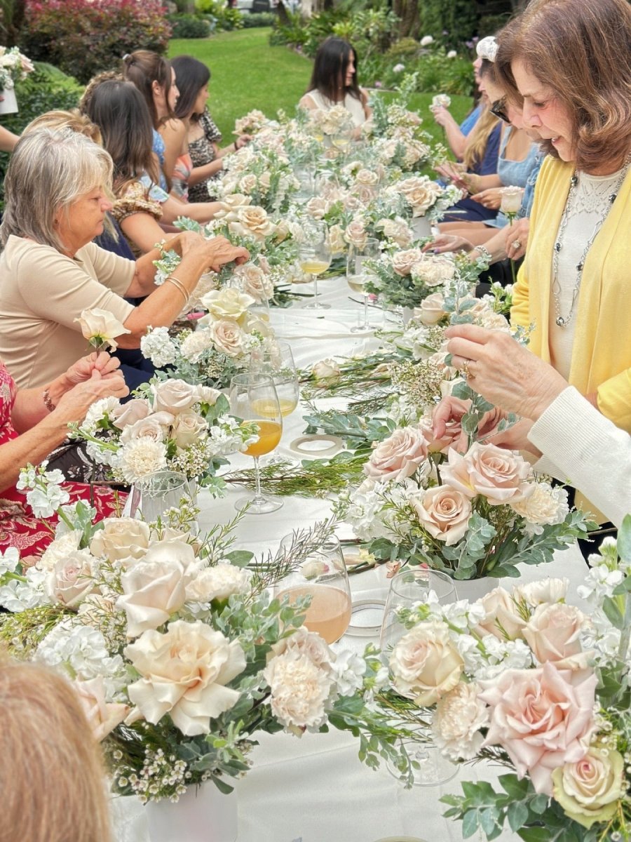 Guests at a long garden table arranging cream and blush roses with eucalyptus, glasses of mimosa between the blooms.