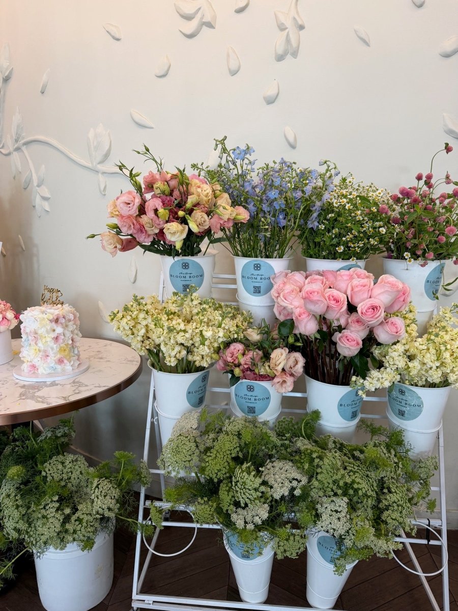 Tiered white flower bar of Bloom Boom buckets — pink roses, blue delphinium, cream stock, and chamomile.